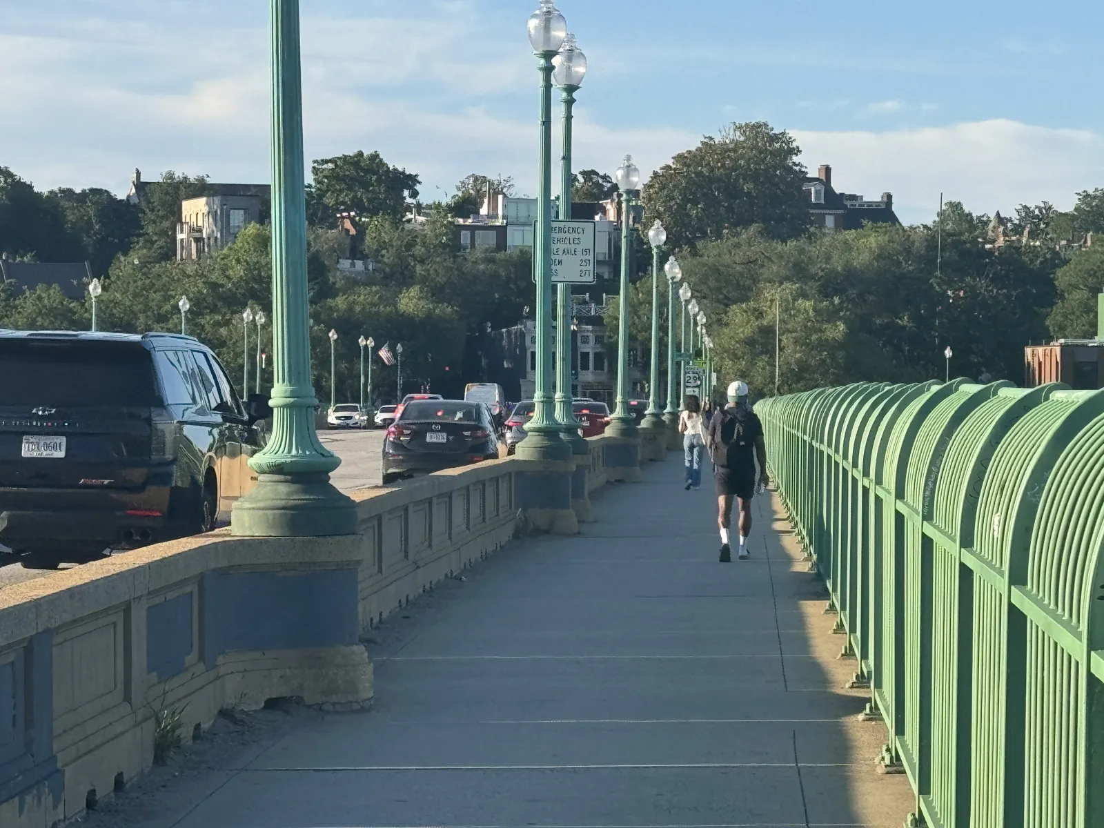 The bridge sidewalk today: the concrete barrier wall is bare gray, alongside the green railings and lampposts