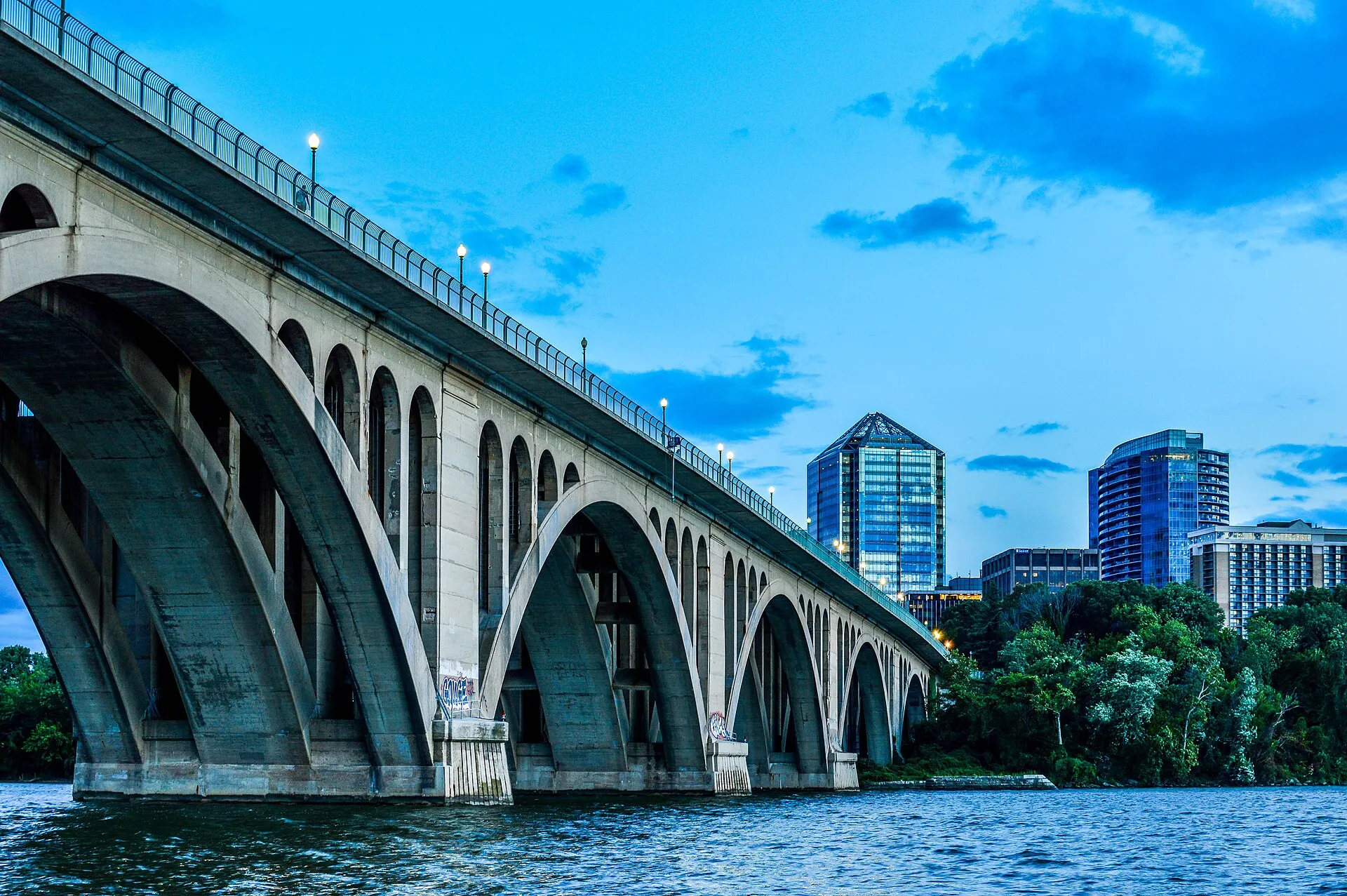 The Francis Scott Key Bridge from below, its concrete arches rising toward the Rosslyn skyline, with graffiti visible on the barrier wall